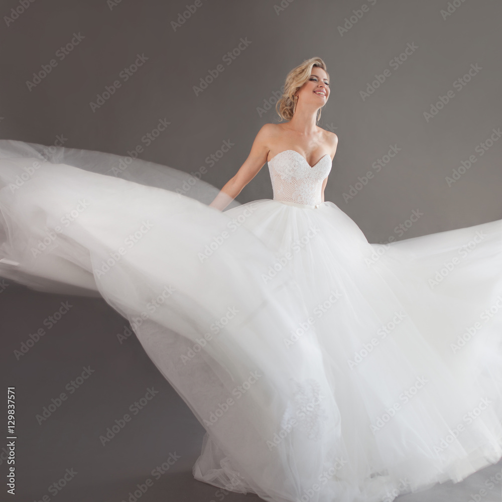 Fototapeta premium Dancing young bride in luxurious wedding dress. Pretty girl in white. Emotions of happiness, laughter and smile, gray background