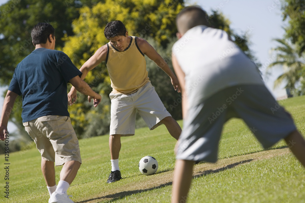 Obraz premium Boy (13-15) with two young men playing soccer outdoors in park.