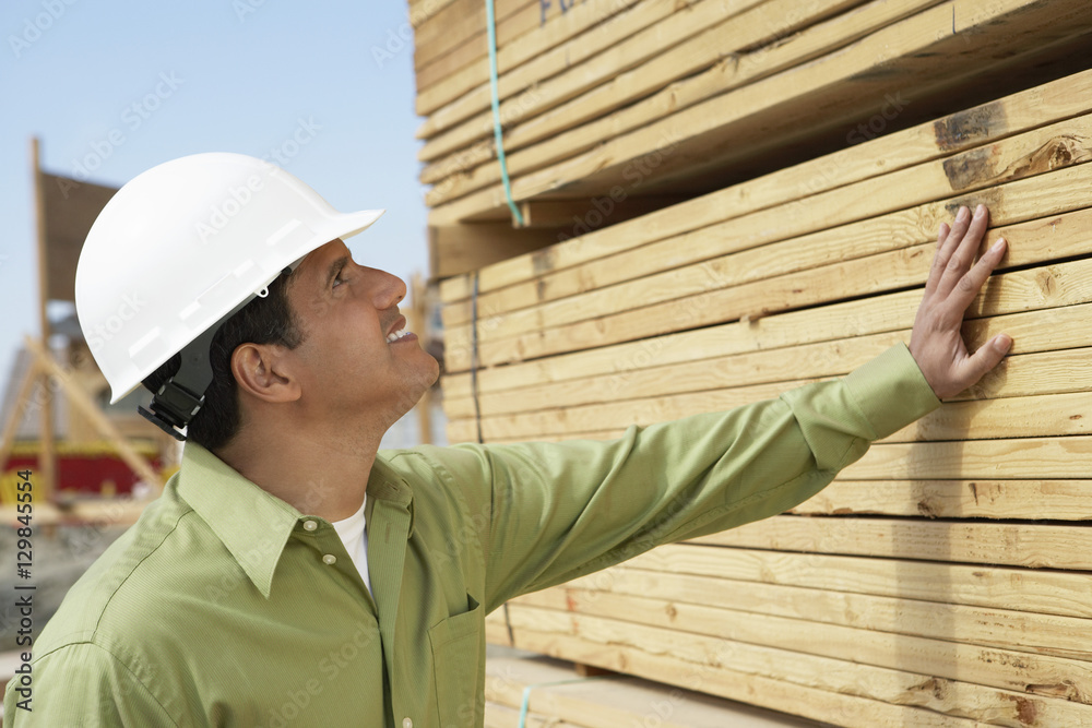 Side view of a smiling construction worker in hardhat inspecting lumber on job site Side view of a smiling construction worker in hardhat inspecting lumber on job site