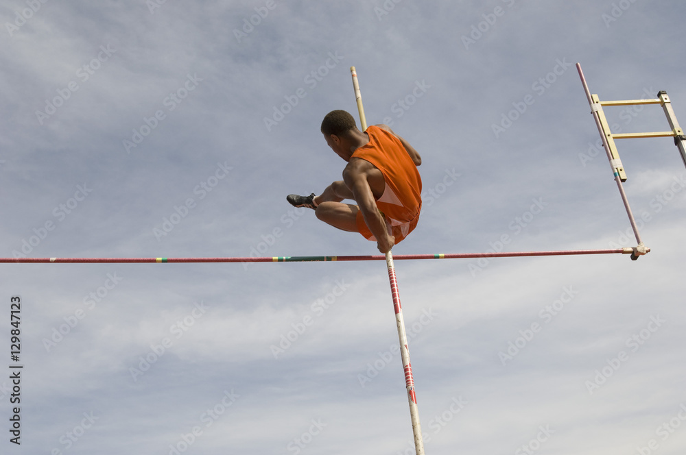 Low angle view of male pole vaulter clearing bar against cloudy sky - obrazy, fototapety, plakaty 