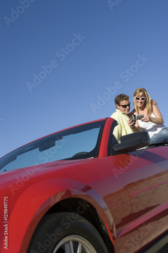 Low angle view of couple sitting on car with handycam against clear sky © moodboard