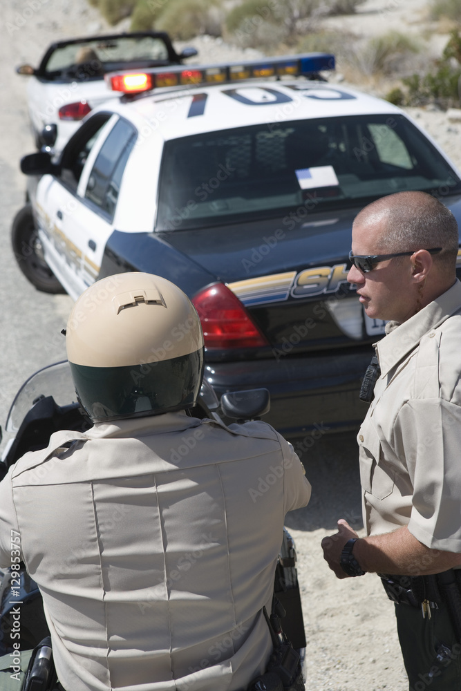 Mature traffic cop looking at coworker sitting on motorbike Mature traffic cop looking at coworker sitting on motorbike