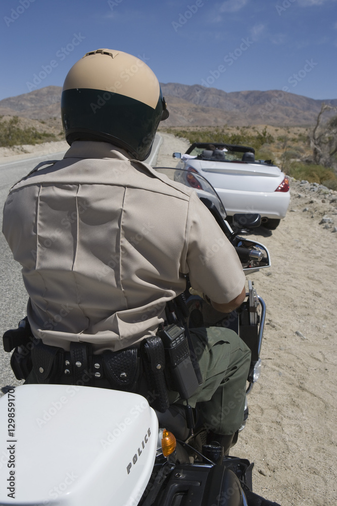 Rear view of a mature traffic cop riding motorcycle with car in the background Rear view of a mature traffic cop riding motorcycle with car in the background
