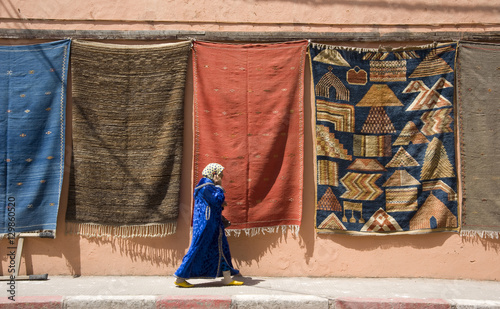 A woman in Islamic dress walking by a wall hung with carpets in the Medina in Marrakech, Morocco