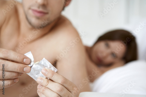 Young man opening condom with woman in bed