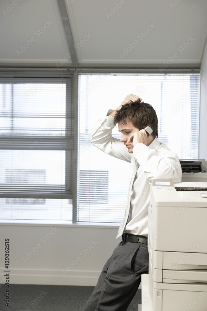Side view of an uncertain businessman leaning on office photocopier and using cellphone
