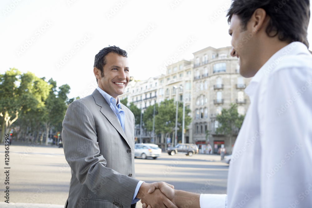 Happy young businessmen shaking hands while standing on city street Happy young businessmen shaking hands while standing on city street