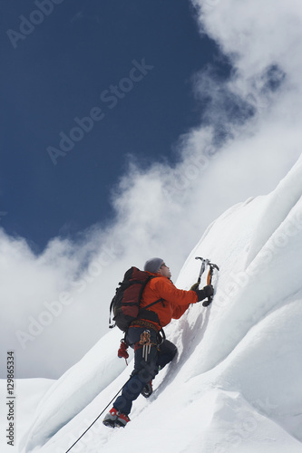 Low angle view of a male mountain climber going up snowy slope with axes against clouds
