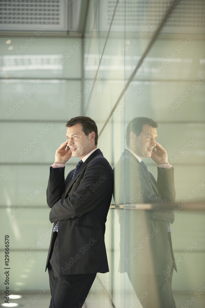 Side view of middle aged businessman using cellphone while leaning on glass wall in office Side view of middle aged businessman using cellphone while leaning on glass wall in office