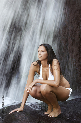 Full length of thoughtful young woman crouching on rock while looking away by waterfall