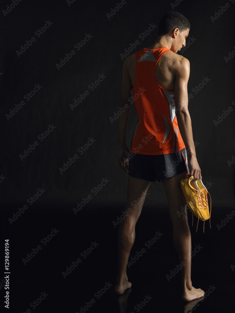 Full length rear view of a male athlete holding shoes against black background
