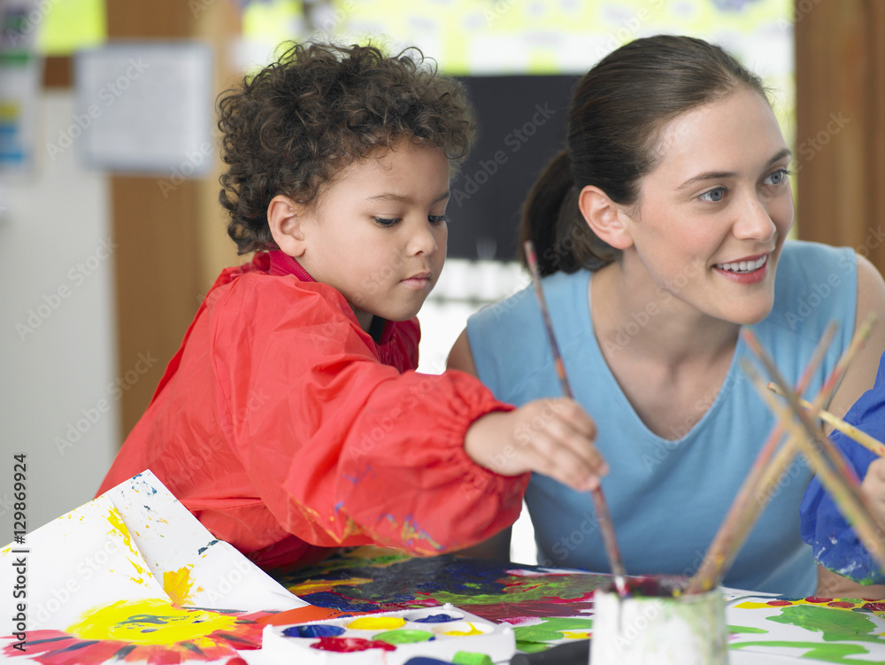 Young boy painting beside teacher in art class