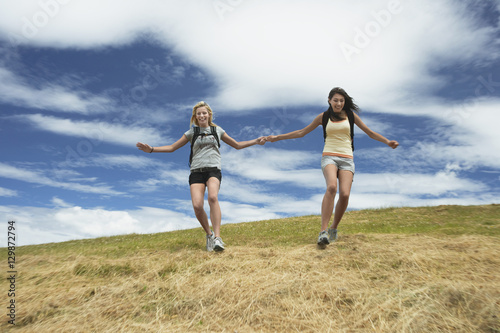 Two women with backpacks holding hands and skipping down hill