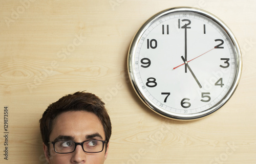 Photography Young businessman wearing spectacles looking at clock on wooden wall in office
