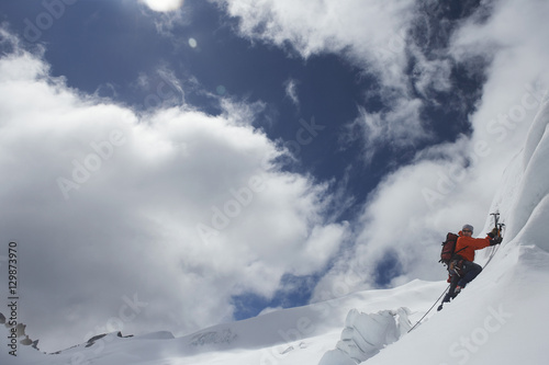 Side view of a male mountain climber going up snowy slope with axes against clouds