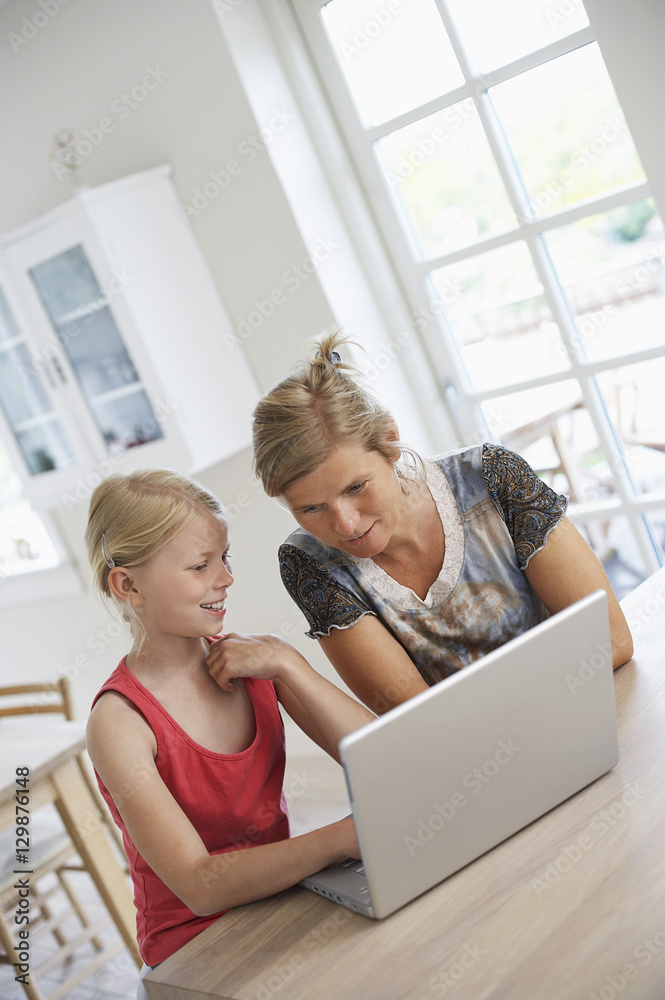 Mother using laptop with daughter at kitchen table Mother using laptop with daughter at kitchen table