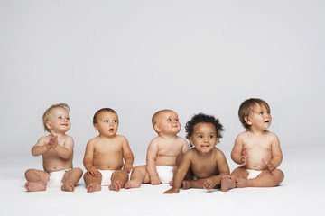 Row of multiethnic babies sitting side by side looking away isolated on gray background