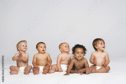Fotografia Row of multiethnic babies sitting side by side looking away isolated on gray bac
