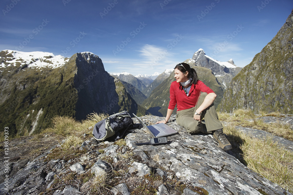 Young woman sitting with laptop on boulder against mountains
