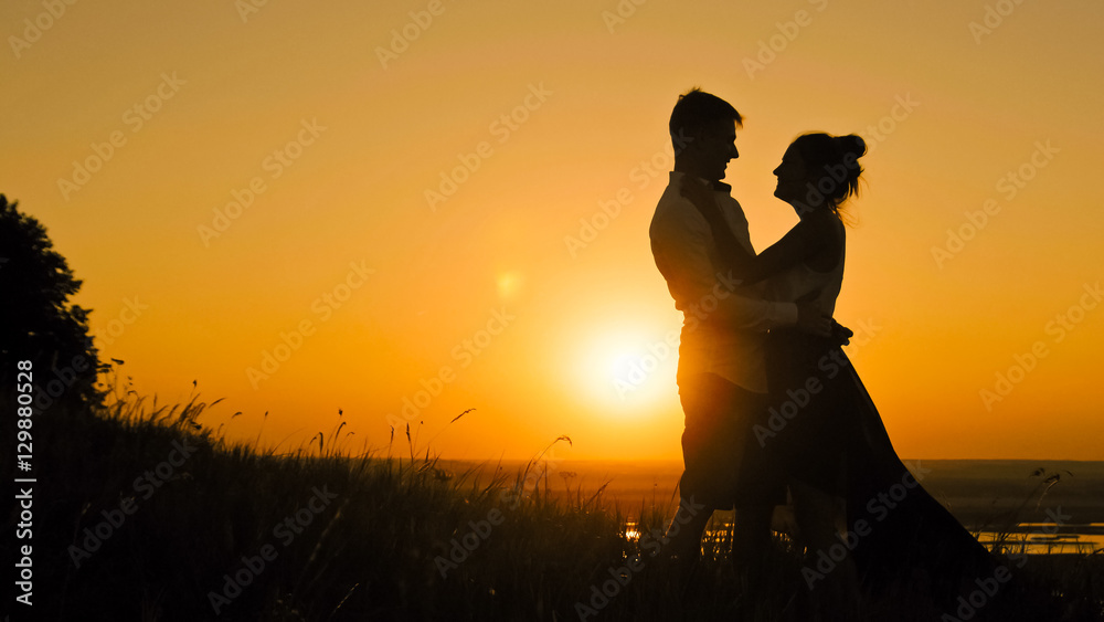 Young loving couple -  man and beautiful girl stands on high hill at sunset  have hugs  kiss, silhouette