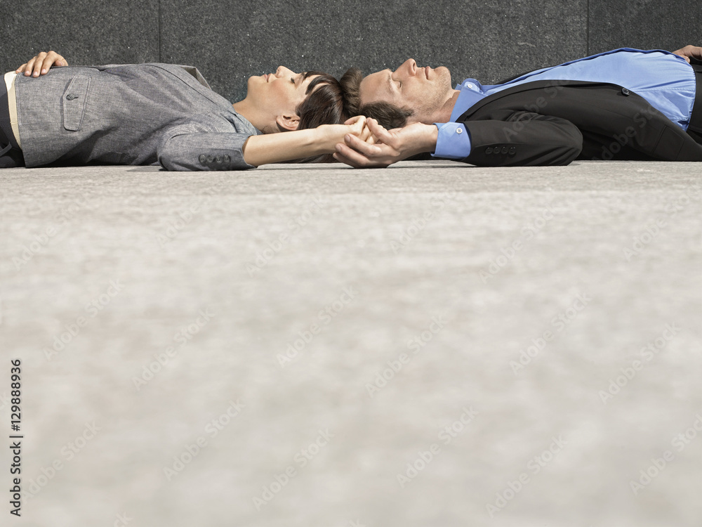 Side view of young businessman and businesswoman holding hands while lying on ground - obrazy, fototapety, plakaty 