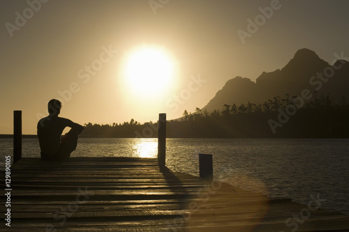 Rear view of man sitting on dock by lake enjoying sunset