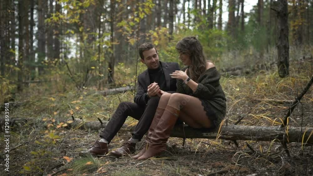 Close up of happy couple having a date in the forest. Smiling man and woman are sitting on the log among the trees. Bearded man in black roll-neck sweater is lovingly looking at his woman