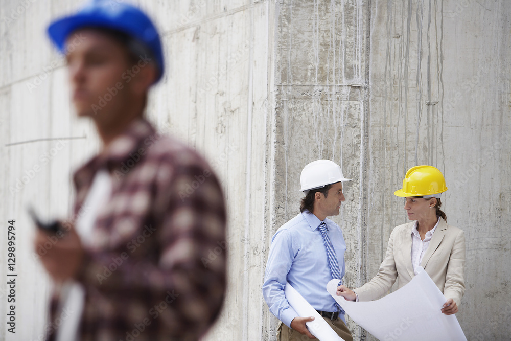 Managers with blueprint and blurred worker in foreground at construction site Managers with blueprint and blurred worker in foreground at construction site