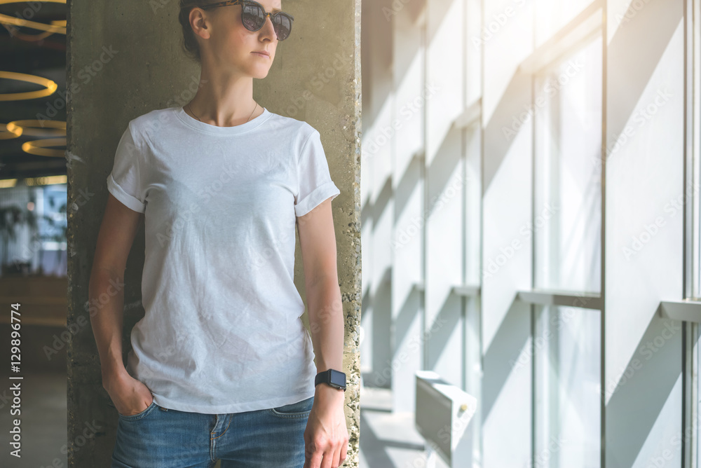 Front view. Young woman in sunglasses, white t-shirt and jeans standing near a window in a room with a modern interior, with his hand in the pocket of jeans. In the background concrete wall.Mock up. Front view. Young woman in sunglasses, white t-shirt and jeans standing near a window in a room with a modern interior, with his hand in the pocket of jeans. In the background concrete wall.Mock up.