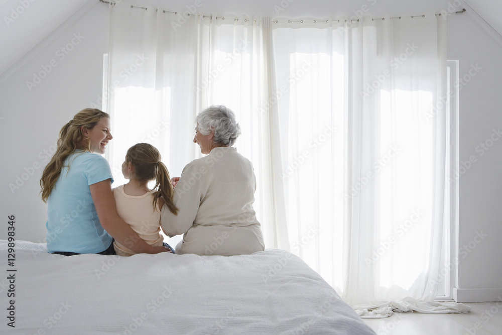 Rear view of girl with grandmother and mother sitting together on bed Rear view of girl with grandmother and mother sitting together on bed