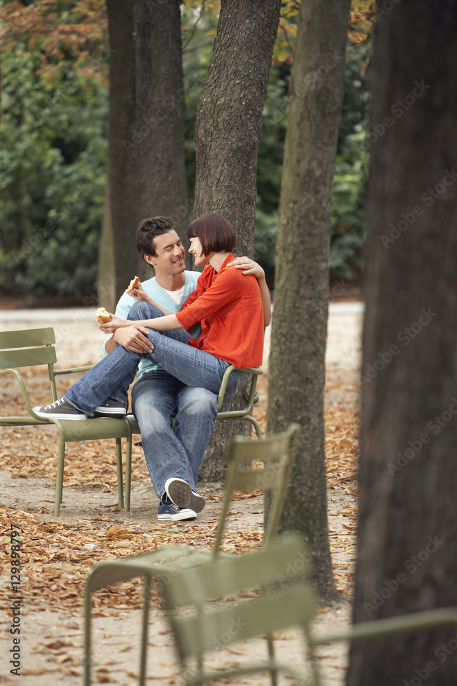 Woman sitting on man while eating snack at the park along tree trunks