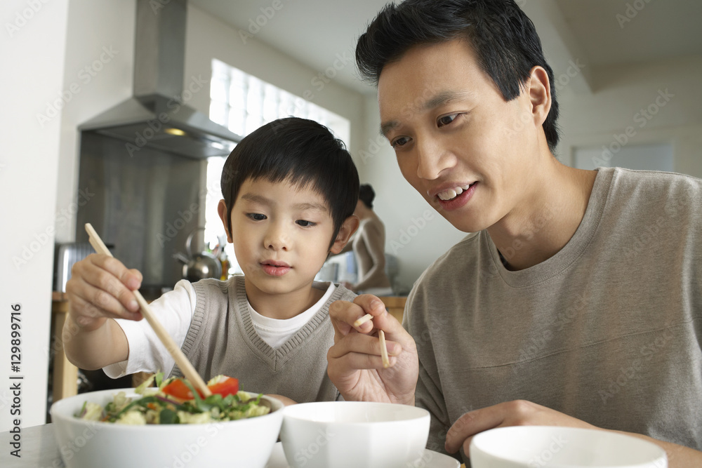 Young father watching son trying to use chopsticks at dining table
