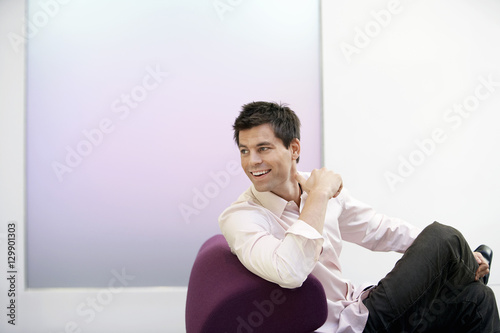 Smiling young businessman sitting on modern sofa with legs crossed looking over shoulder
