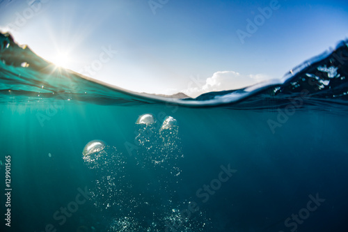 Ocean water line splits sky and underwaer part. Air bubbles in deep blue. Bright sun light and blue clear sky