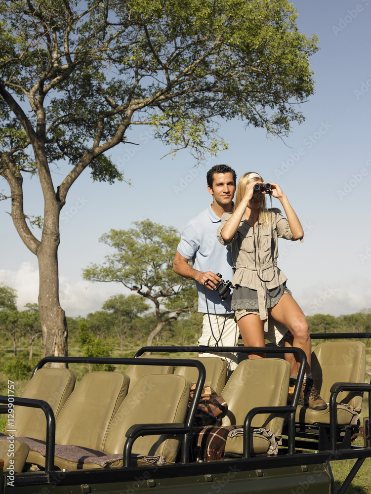 Obraz premium Young couple on safari standing in jeep and looking through binoculars