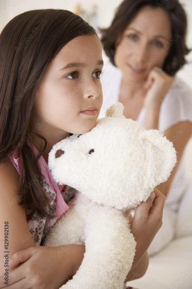 Sad young girl hugging teddy bear with mother in background Sad young girl hugging teddy bear with mother in background