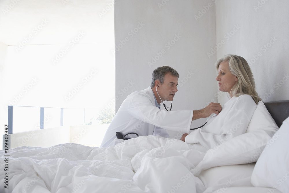 Middle aged doctor examining woman with stethoscope in bed at home