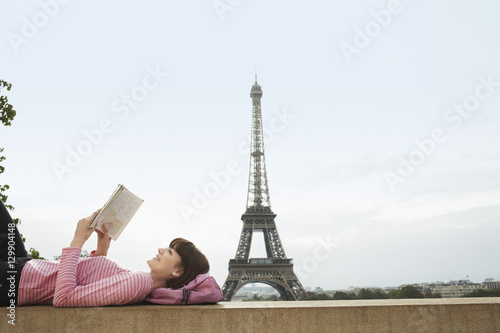 Photography Side view of a young woman lying on balcony and reading book in front of Eiffel