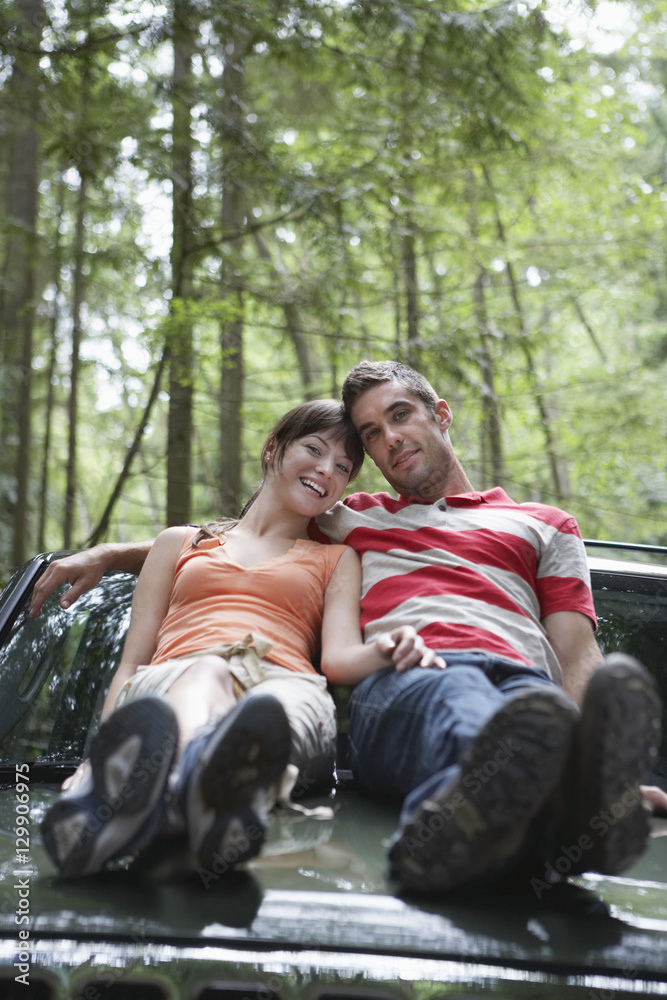 Full length portrait of a smiling young couple sitting on car bonnet in forest Full length portrait of a smiling young couple sitting on car bonnet in forest