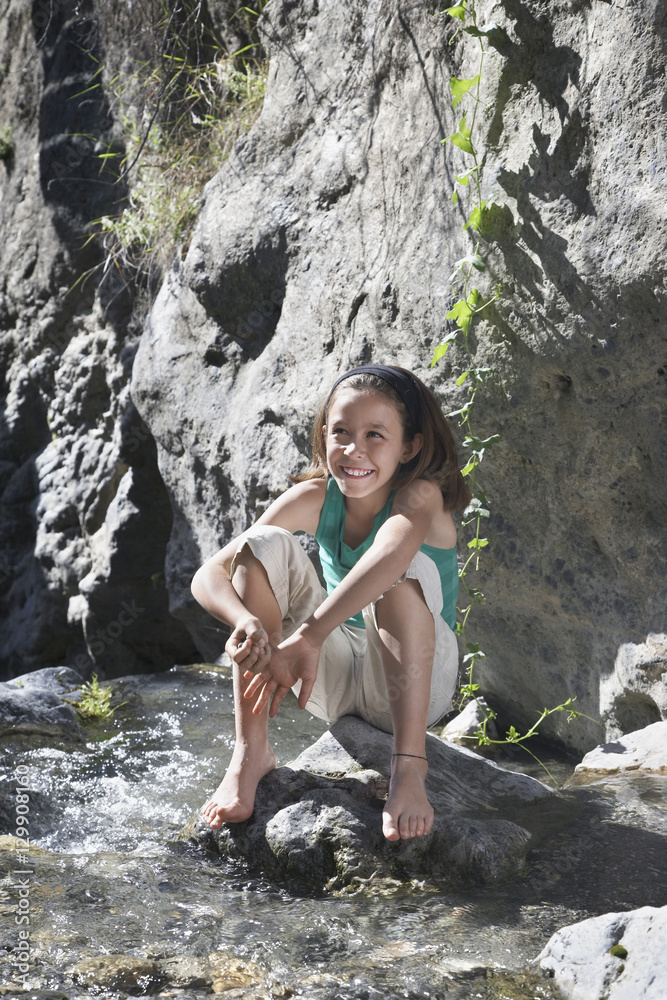 Full length portrait of a smiling little girl sitting on rock by stream Full length portrait of a smiling little girl sitting on rock by stream
