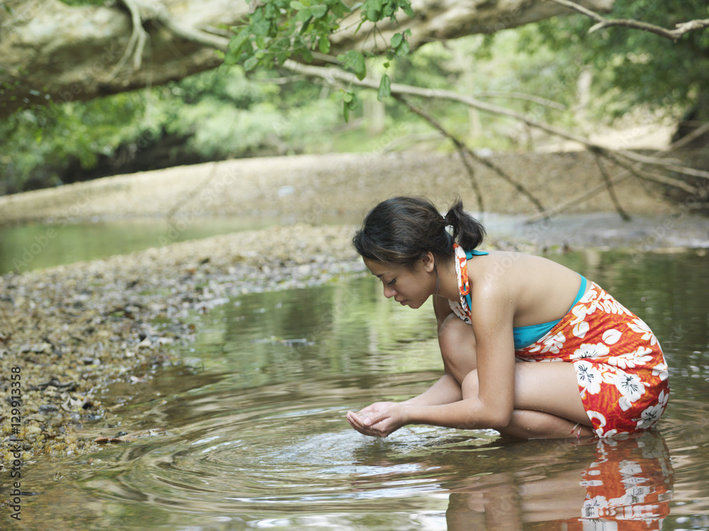 Fototapeta premium Side view of a young mixed race woman squatting in forest lake with hands cupped