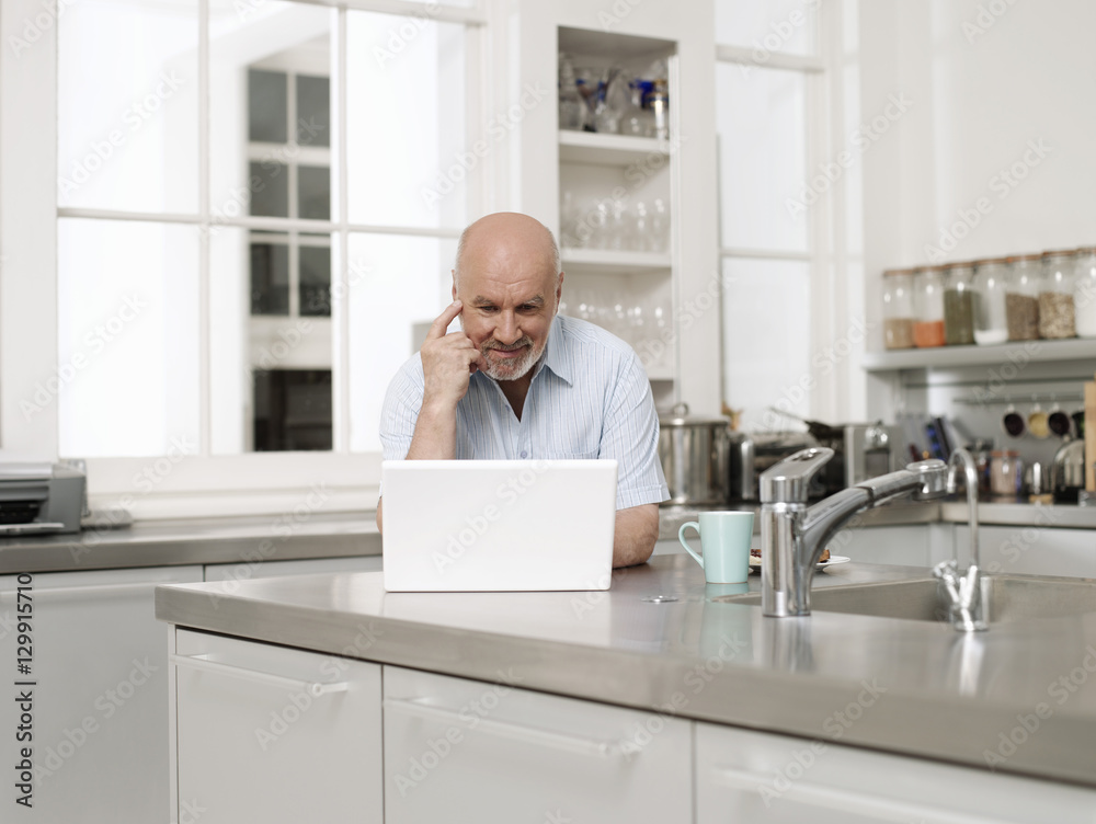 Smiling mature man using laptop in kitchen at home Smiling mature man using laptop in kitchen at home