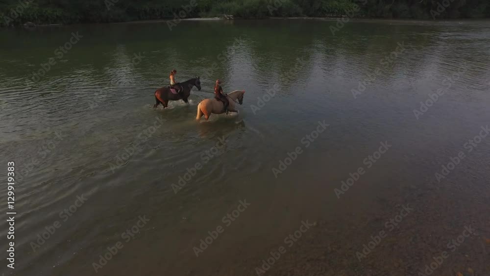 AERIAL: Two female riders riding horses in wide river at late sunny morning AERIAL: Two female riders riding horses in wide river at late sunny morning