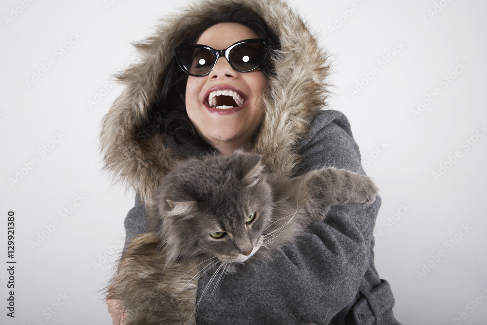 Laughing young woman in hooded fur coat holding cat on gray background