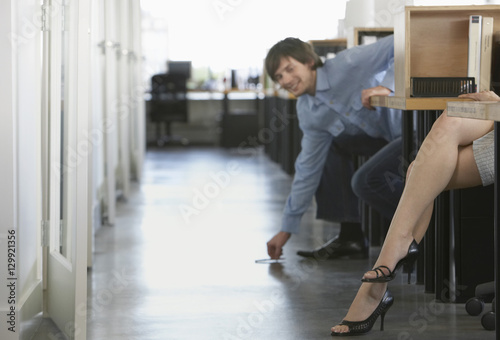 Smiling businessman picking pen off floor near woman's legs in office