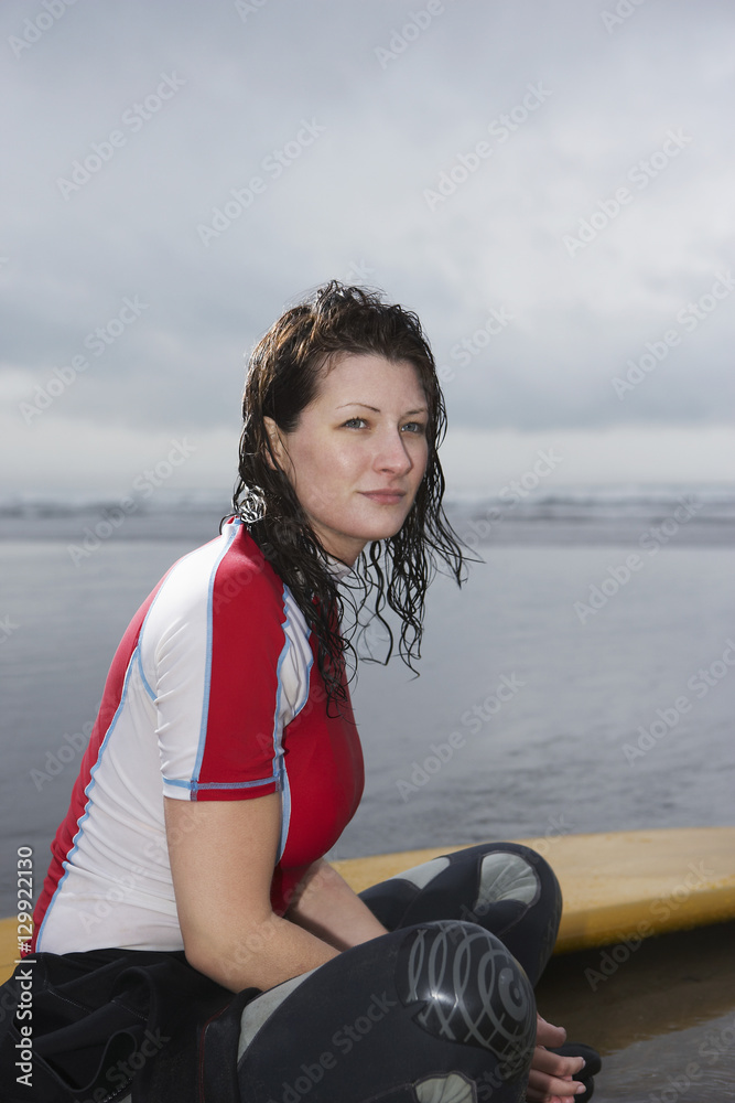 Fototapeta premium Side view of thoughtful young woman with surfboard looking away while sitting on beach