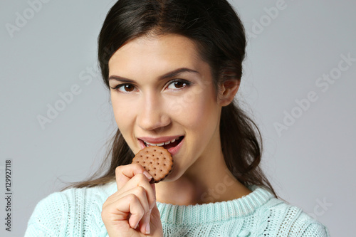 Pretty young woman eating tasty cookie, on grey background