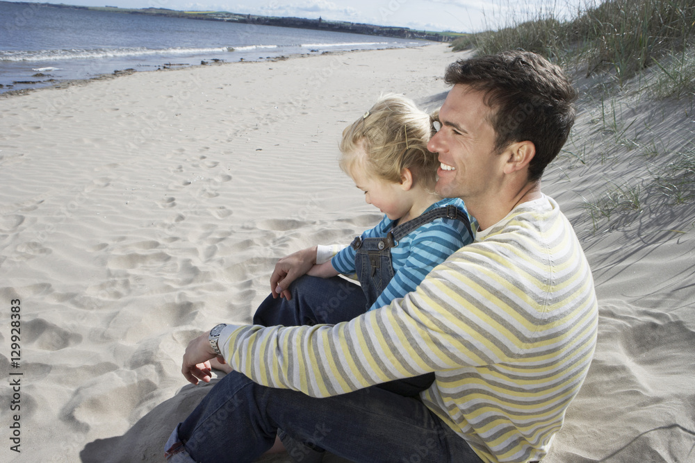 Side view of happy father and daughter sitting on sandy beach