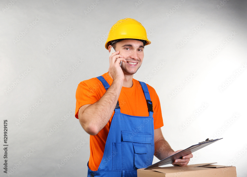 Handsome warehouse worker talking by mobile phone while standing near carton box, on light background