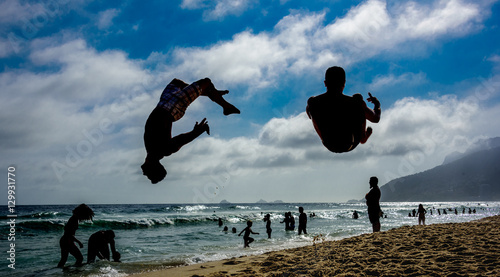 Silhouettes of two men performing somersaults at sand of Ipanema beach with silhouettes of swimming people, blue sky, white clouds and silhouettes of mountains at background, Rio de Janeiro, Brazil